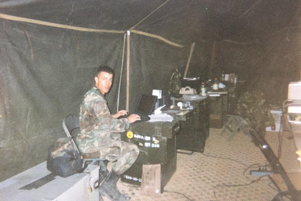 Rick Rivetti on deployment with the Marines, working on a laptop at a field desk inside a tent
